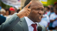 Ayaba Cho Lucas speaking into a microphone with his index finger raised.
