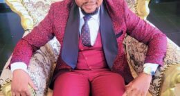 Dr Success Nkongho seated in an ornate chair, wearing a red patterned suit.