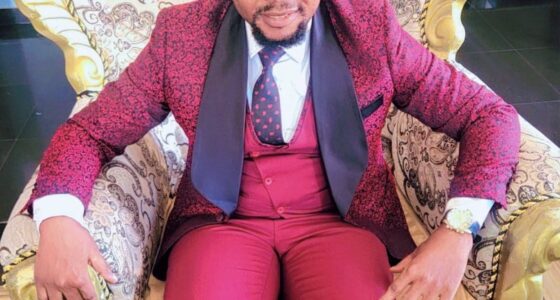 Dr Success Nkongho seated in an ornate chair, wearing a red patterned suit.