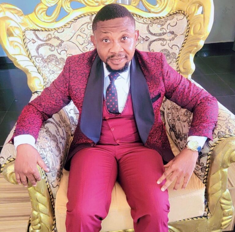 Dr Success Nkongho seated in an ornate chair, wearing a red patterned suit.
