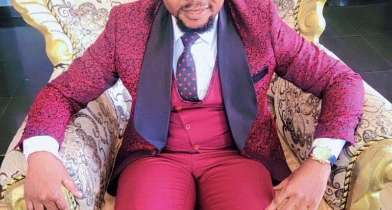 Dr Success Nkongho seated in an ornate chair, wearing a red patterned suit.